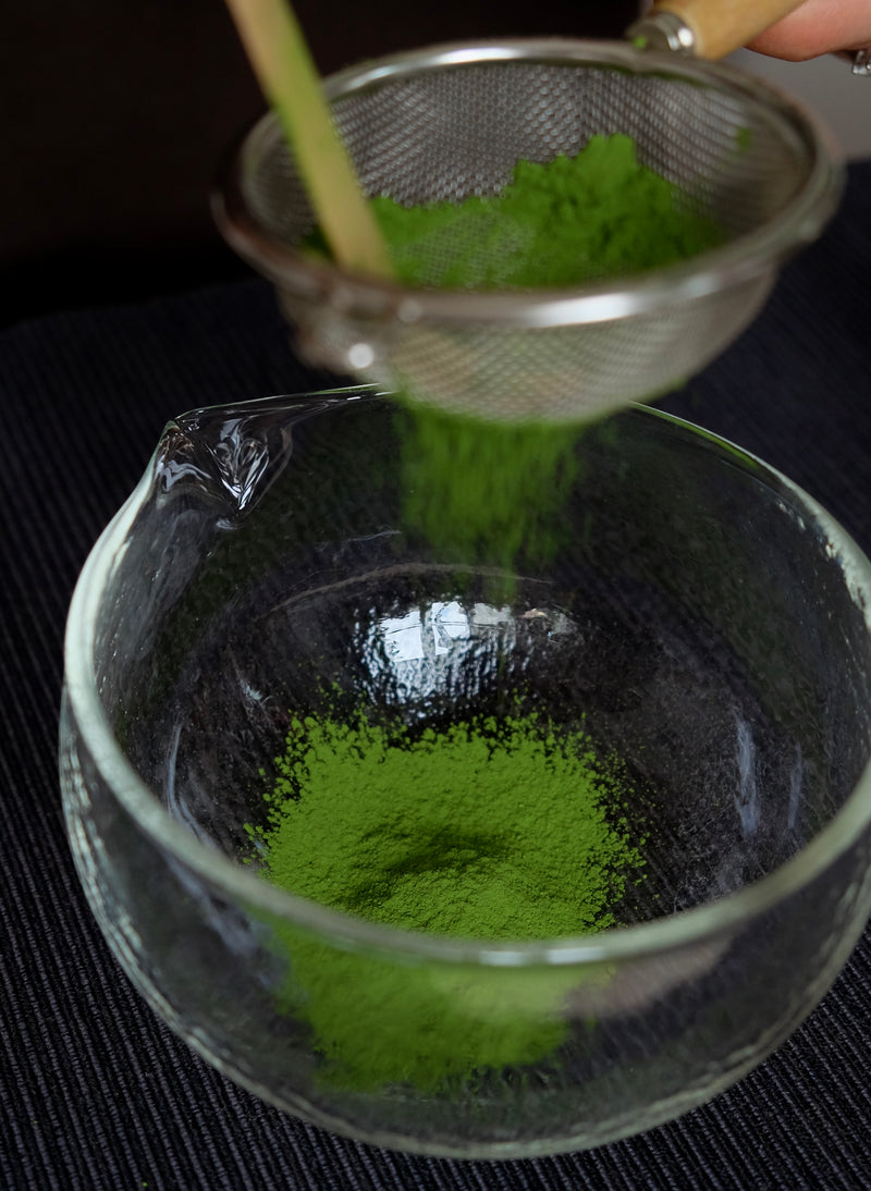 Green powder being sifted into a glass bowl using a metal strainer.