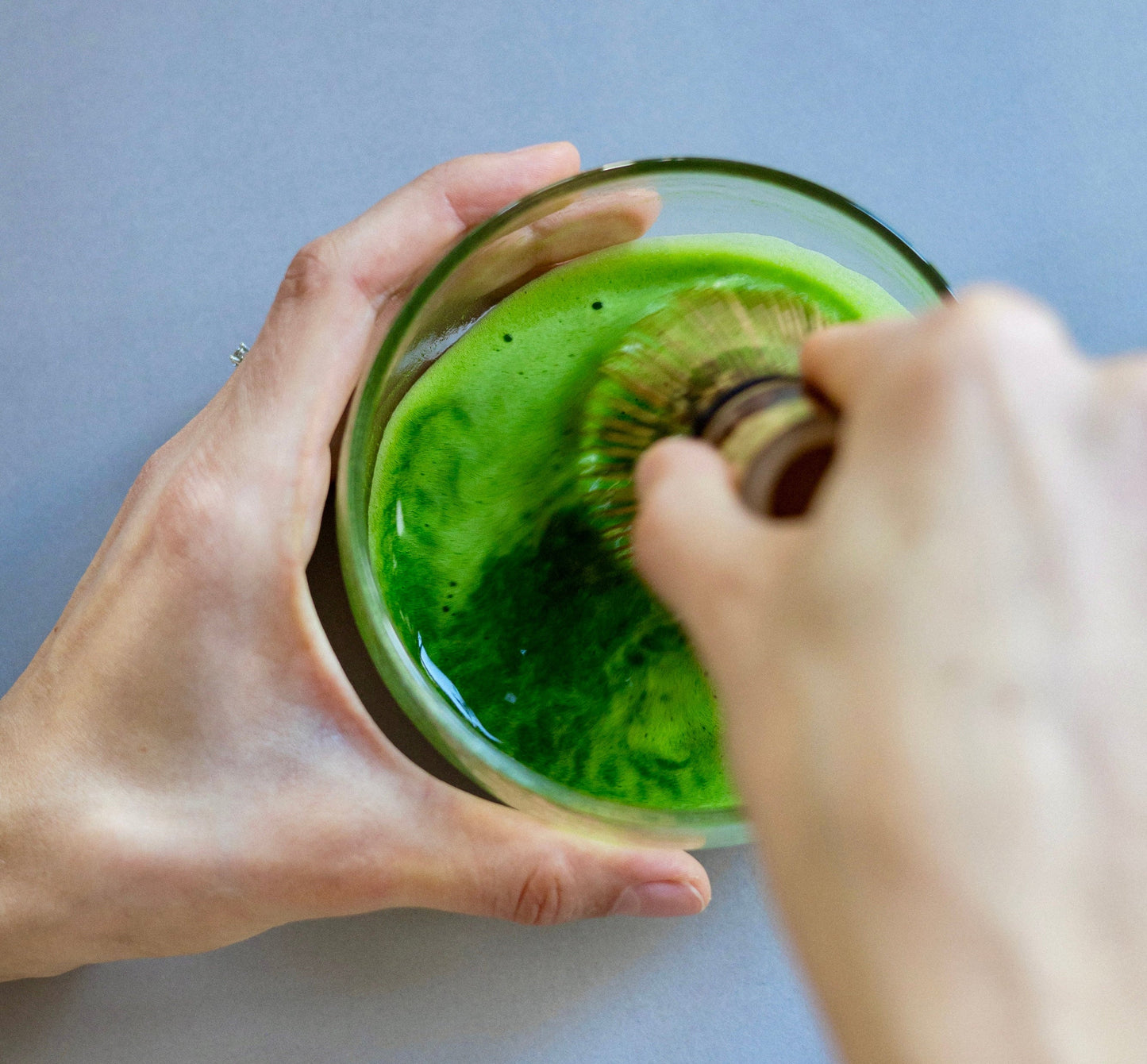 Person stirring a matcha in a glass bowl with a whisk on a light blue background