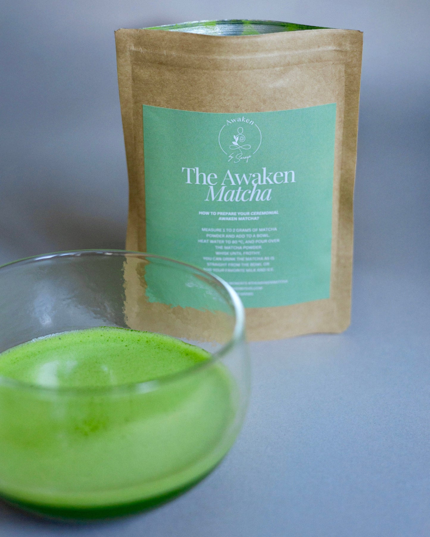 Glass of green matcha tea with a package labeled 'The Awaken Matcha' on a gray background