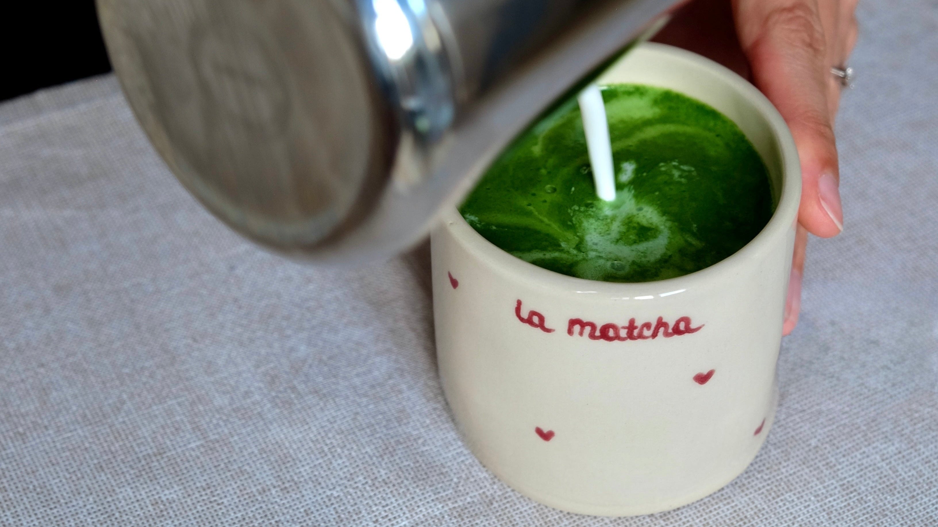 Person pouring a matcha  into a cup labeled 'La Matcha' on a textured surface.