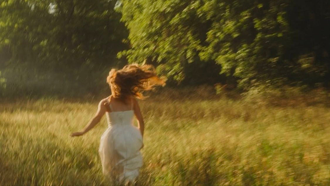 Woman in a white dress running through a grassy field with trees in the background
