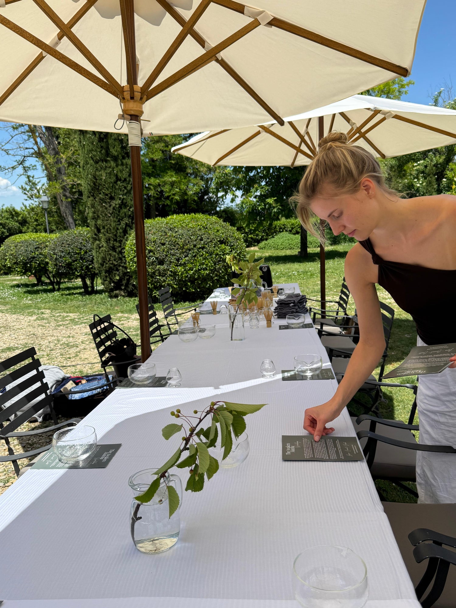 Person setting up a table outdoors with white tablecloth, glasses, and greenery.